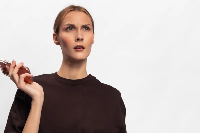 Confused young woman with light hair holds smartphone in hand, looks puzzled and surprised, isolated on white background. Concept of misunderstanding, doubt, and communication issues.