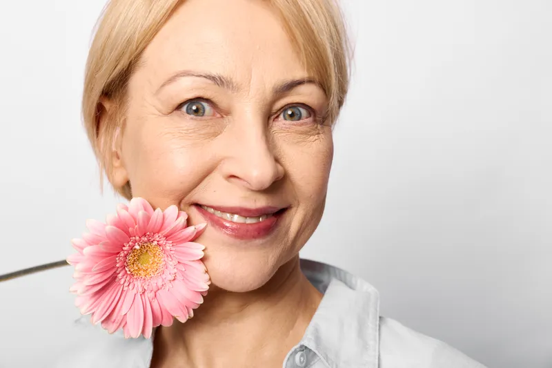 Woman elderly smiling holding pink flower closeup portrait, soft light background, calm joyful expression, beauty skincare concept, gentle care atmosphere.