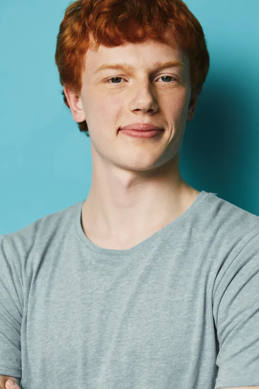 Redhead young man portrait with freckles wearing gray t shirt standing against blue background looking at camera with slight smile. Casual style and natural light for people concept.