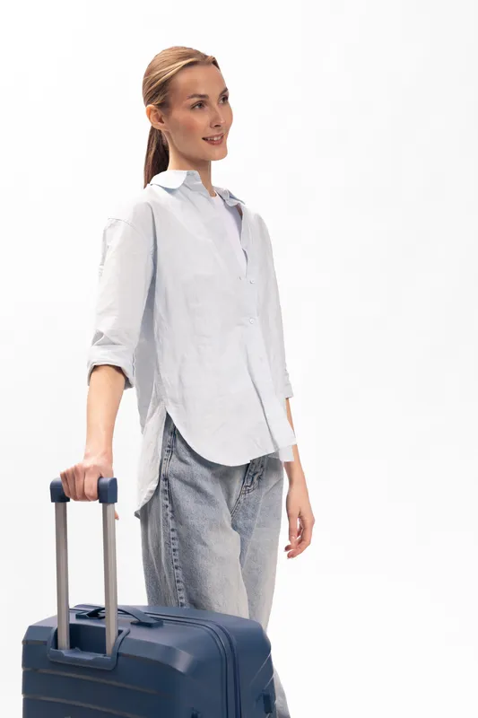 Young woman in casual clothes stands with blue suitcase on white background. Concept of travel, tourism, and vacation preparation with relaxed and confident mood.