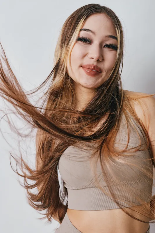 Young woman with long hair wearing beige top smiling and posing with hair flowing in front of white background. Beauty, casual, confident and joyful portrait in studio.