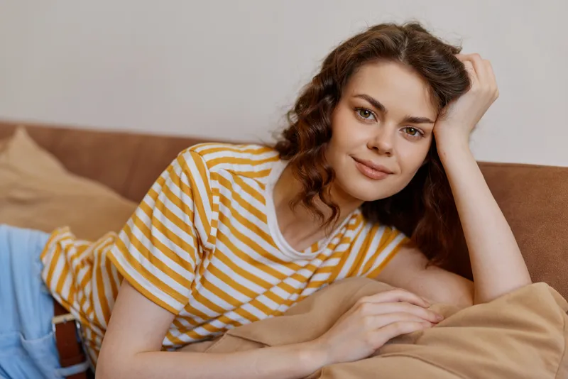 A relaxed woman lounges on a sofa in a striped tee and jeans, resting her head on a hand. A warm, casual home scene conveying comfort, calm, and everyday life.