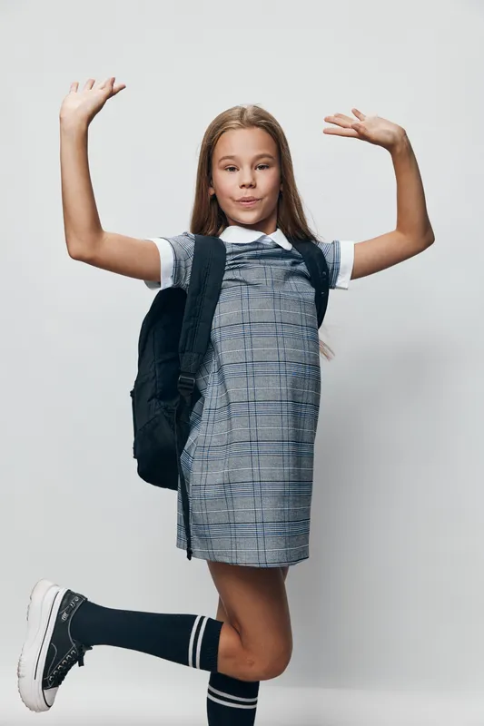Girl teenager with backpack in casual dress and knee socks posing with hands up on white background. Studio portrait of young student ready for school or activity.