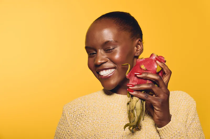 Smiling African woman with dark skin holds exotic fruit near face, wearing knitted sweater on yellow background. Concept for freshness, health, natural food, and vibrant lifestyle.
