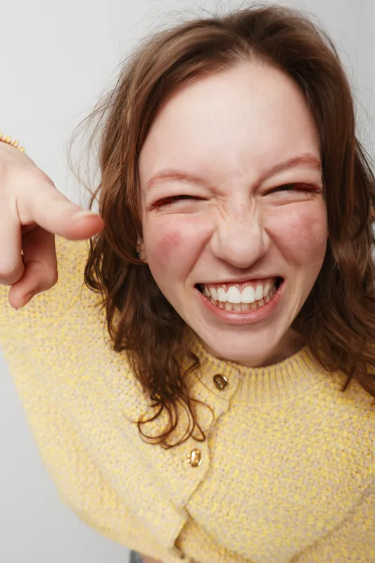 Happy young woman with curly hair wearing yellow sweater smiles widely and points finger at camera. Close-up portrait on light background. Concept of joy, fun, and positive emotions.