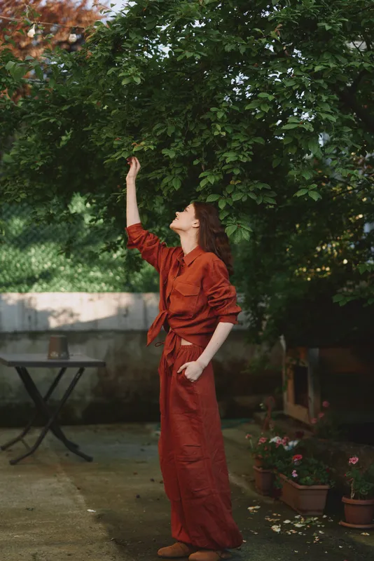 Woman outdoors in a red jumpsuit reaches toward leaves, a sunlit fashion portrait with relaxed posture, natural colors, and lush greenery in the background