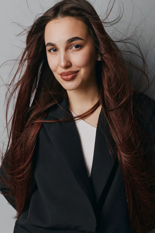Young woman with long hair wearing black blazer and white top posing with confident smile and wind blowing hair on gray background. Portrait of attractive female with natural makeup and earrings.