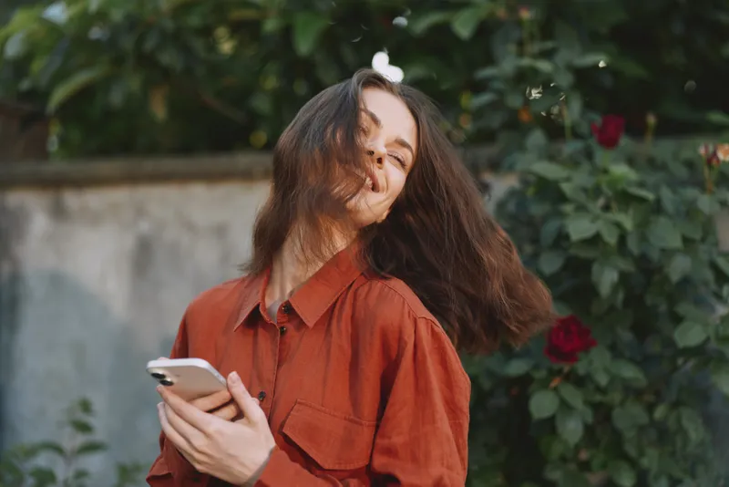 A woman in a red shirt enjoys a sunny outdoor moment, smiling with a phone in hand among blooming roses, capturing casual everyday life and carefree joy