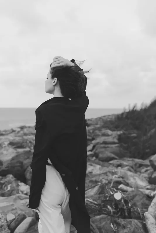 Woman wind coast sea jacket landscape, moody outdoor scene with a solitary figure on a rocky shore, wind in hair, looking toward the horizon by the sea, dramatic yet serene travel style