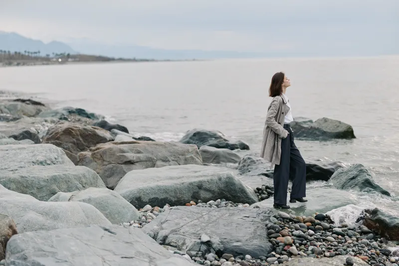 A woman in casual clothes stands on rocky shore looking at the sea, enjoying a peaceful moment. The cloudy sky and calm water create a serene coastal atmosphere.