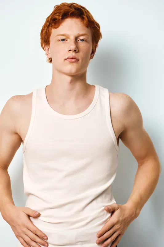 Young man with red hair wearing white tank top stands against white wall. Casual style, confident pose and natural light highlight his clean skin and serious expression on face.