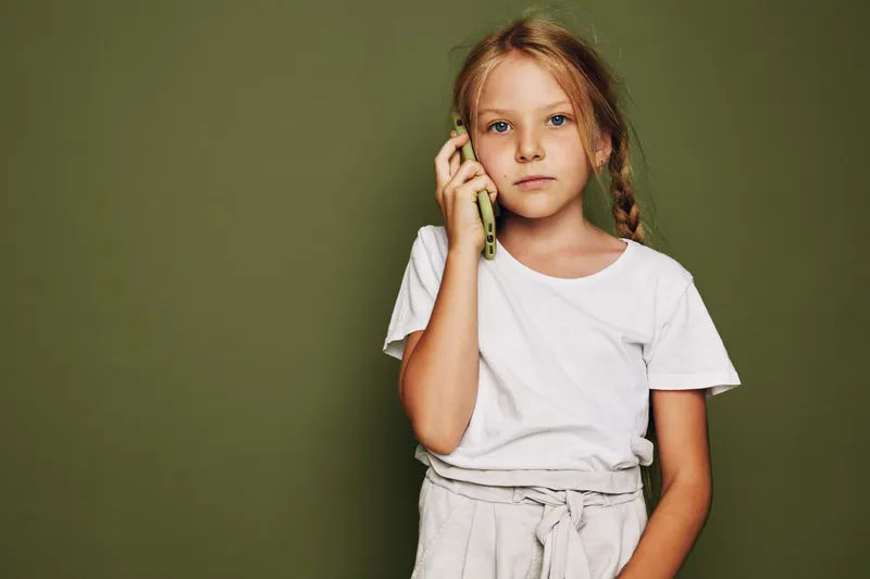 Serious young girl with braided hair wearing white t-shirt holds a phone to her ear standing against a plain green background. Concept of communication and childhood.