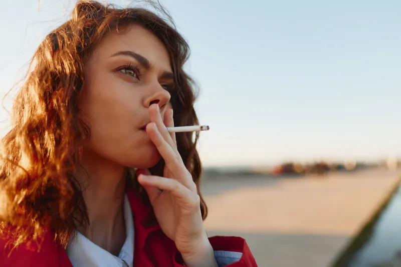A young woman with curly hair smokes a cigarette outdoors during daytime, looking thoughtful. The background shows a blurred urban setting under clear sky.