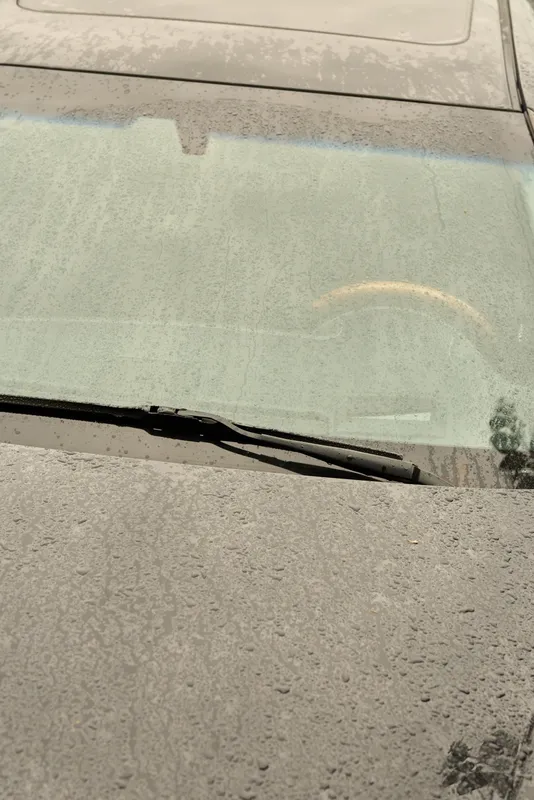 Rainy urban car scene showing a windshield with a wiper in motion, droplets on glass, muted reflections, and a moody atmosphere of daily commute and gray skies