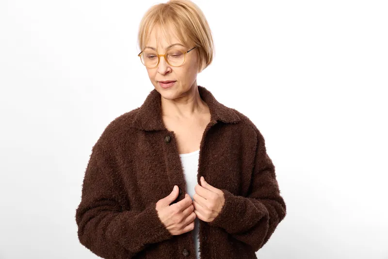 Portrait of elderly woman with glasses in wool cardigan on white studio background, looking down with concerned expression, hands holding sweater front gently