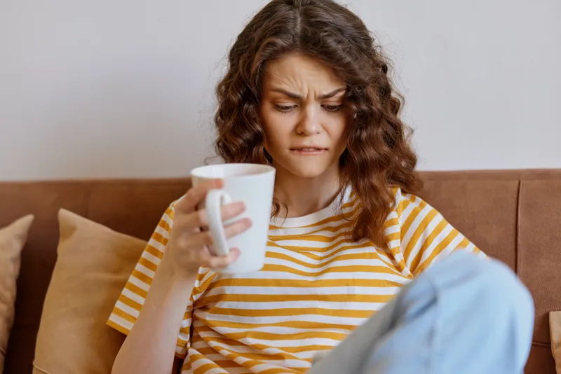 Woman at home sits on a sofa in a striped shirt, holding a mug and frowning, conveying a tired mood during a quiet morning in a casual lifestyle scene.