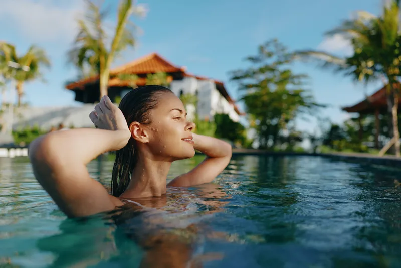 A relaxed woman rests in a bright pool, eyes closed, hands behind head, enjoying warm sunlight. A tropical resort setting with palms frames a calm moment of leisure and wellness.