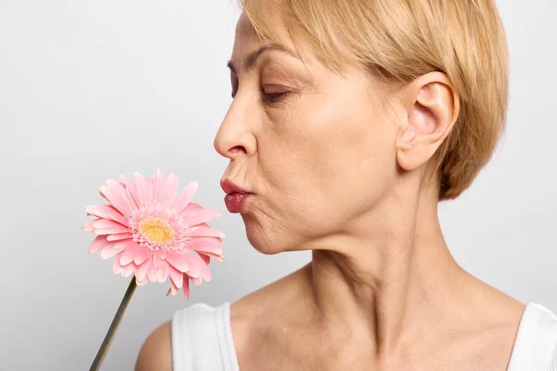 Woman smelling pink gerbera flower, closeup portrait of blonde, calm spring mood, eyes closed, lips near flower, soft light on white background, minimal composition