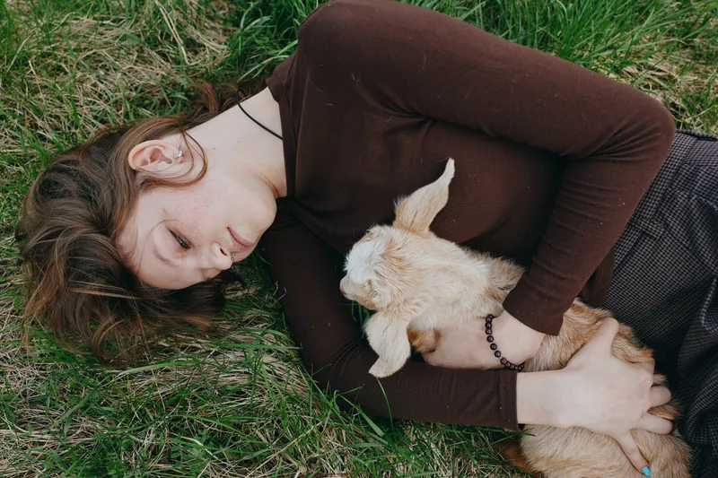 A woman lies on grass in a brown sweater, holding a gentle goat kid. The scene conveys a calm countryside lifestyle, natural bond, and tender animal companionship outdoors.
