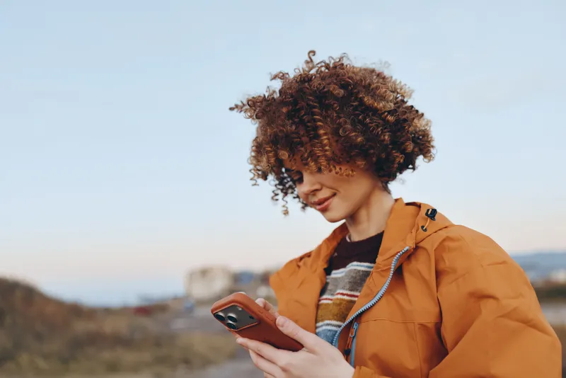 Woman lifestyle outdoors smartphone, curly hair, jacket portrait, mountains and sky, smiling while scrolling on phone, warm sunset light, casual travel mood