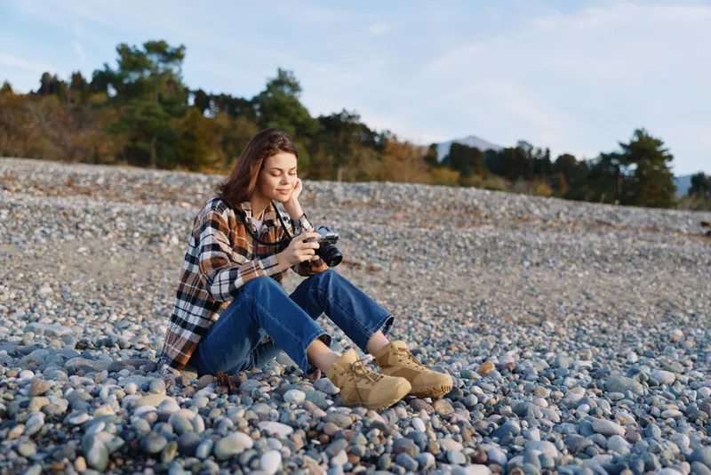 Woman sitting on a pebble beach with a camera, dressed in flannel and boots, enjoying outdoor photography and a calm coastal moment at the quiet shore