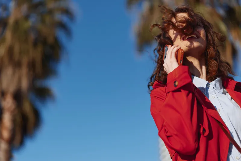 Young woman in red jacket talking on smartphone outdoors with palm trees and clear blue sky in background, smiling and enjoying sunny day in urban setting.