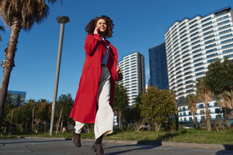 Young woman with curly hair wearing a red coat and white pants walks confidently on a city sidewalk under clear blue sky during daytime.