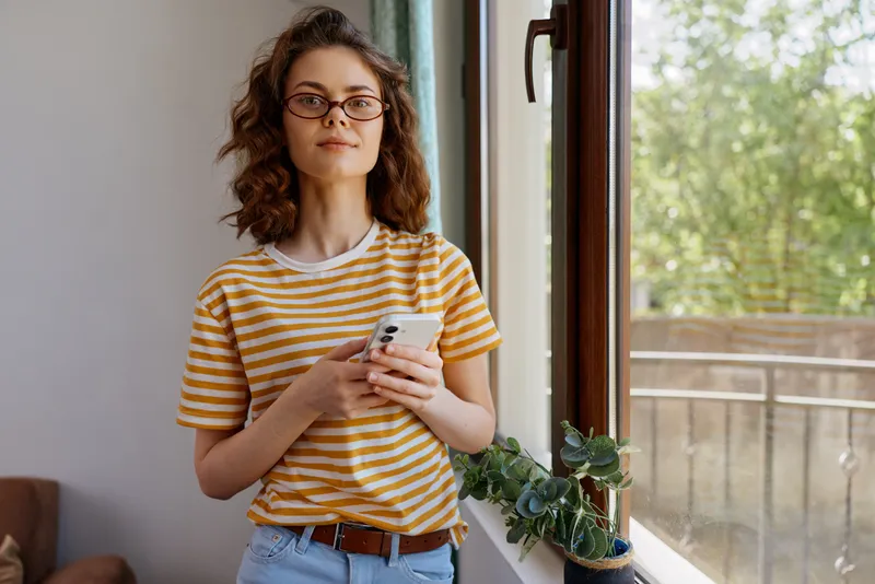 Young woman with curly hair wearing a striped tee stands by a bright window, calmly looking at her phone, embodying a relaxed home moment