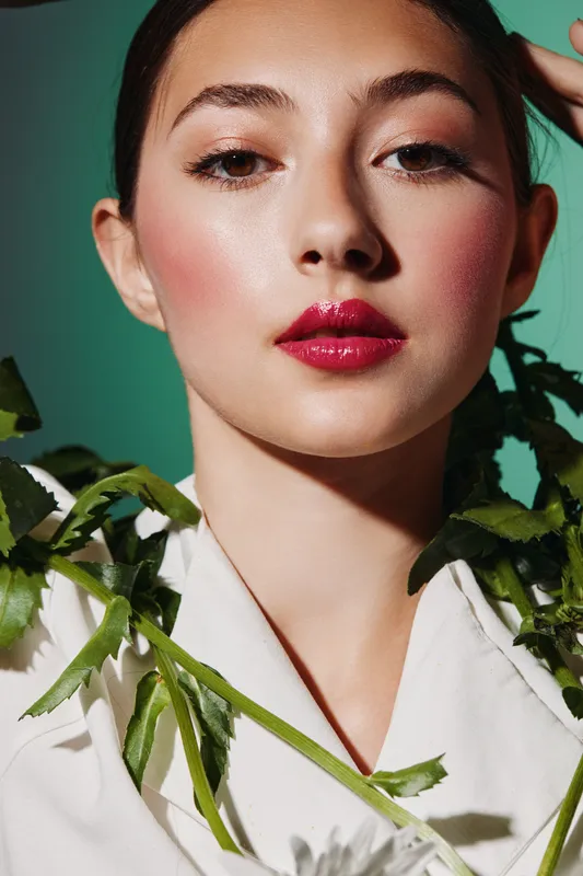 Close-up of a young woman with glossy red lipstick and glowing skin in creative fashion styling with green plants on white outfit. Studio shot beauty concept.