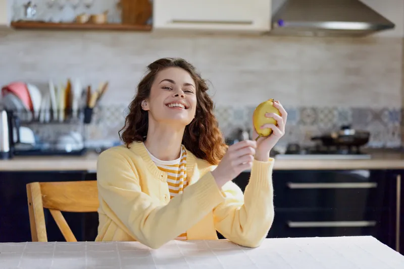 Woman in a yellow cardigan sits at a kitchen table, smiling and holding an apple, a bright cozy home lifestyle moment with natural light and relaxed mood.