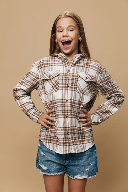 Girl with long hair in plaid shirt and denim shorts stands confident with hands on hips smiling and expressing happiness in studio with beige background and casual style.