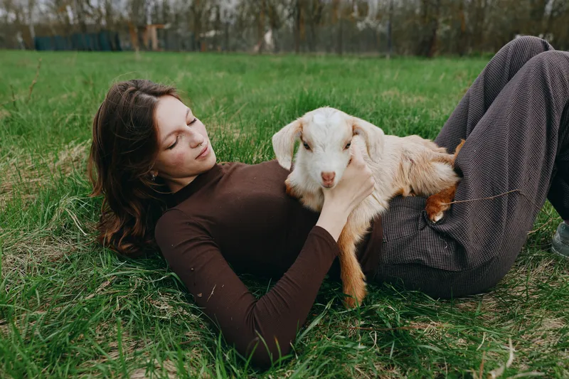 Lifestyle woman with goat kid lying on grass outdoors relax nature friendship calm pasture under warm natural light candid moment of care and connection in a rural setting