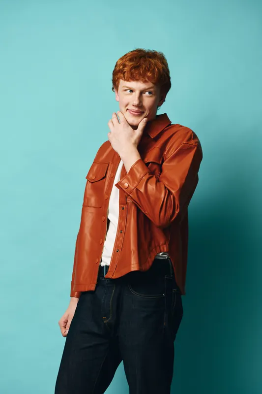 Young man with red hair wearing brown jacket and white shirt posing with hand on chin looking away on blue background. Casual style with thoughtful expression and denim pants.