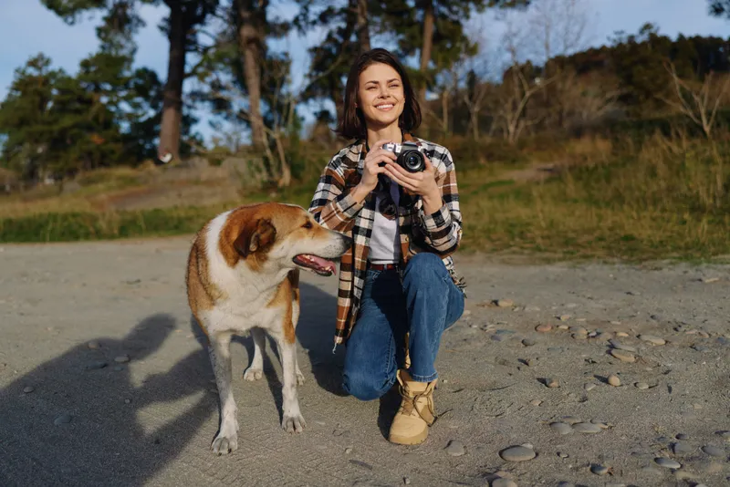 Woman photographer dog outdoors camera nature, a candid moment showing a cheerful dog beside a smiling photographer on a gravel path during outdoor exploration with sunlit trees in background and a
