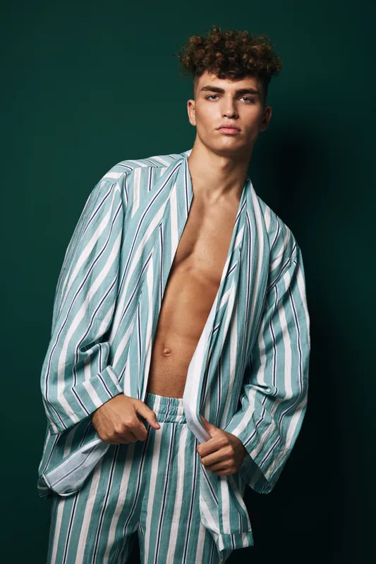 Young man with curly hair wearing striped pajamas standing in front of dark green background. Male model with serious expression posing with casual indoor clothing in studio light.