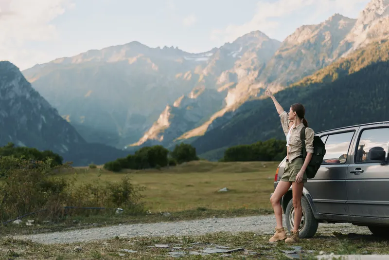 Young woman with a backpack stands beside a parked car in a mountain valley, raising her arm toward the sky as golden light hits rugged peaks, her expression curious and hopeful.