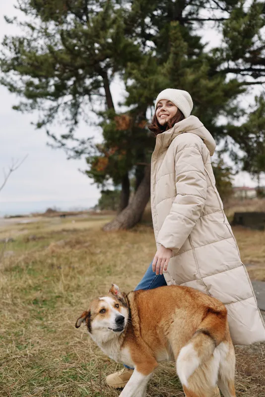 Woman in a puffy beige coat and beanie walks outdoors with a friendly dog on a cool day in a park, trees in the background, relaxed mood, casual fashion and pet companionship