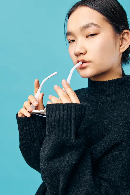 Young woman holding white glasses near mouth wearing black sweater with long sleeves on blue background. Portrait of thoughtful female with clean skin and natural makeup in studio.
