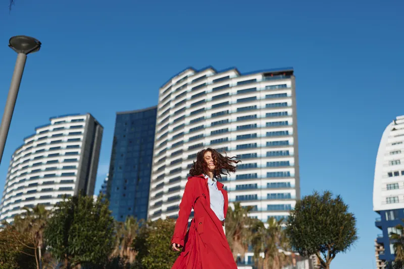 Young woman with curly hair wearing a red coat walks confidently in front of modern high-rise buildings on a sunny day, expressing freedom and urban lifestyle.