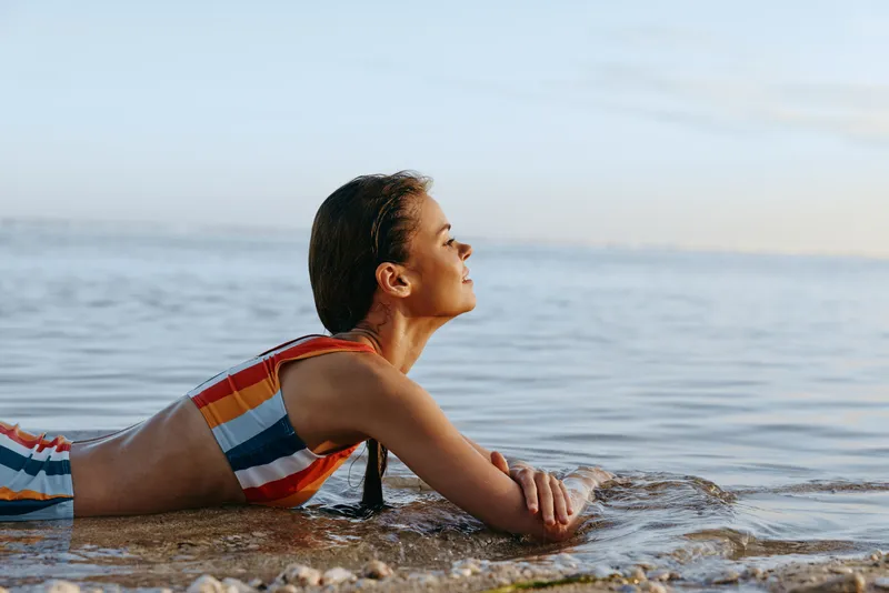 Woman on the beach in shallow ocean water, swimming calmly with gentle sunlit light, relaxed posture, summer day mood, soft waves, natural colors, peaceful seaside horizon, warm breeze smile