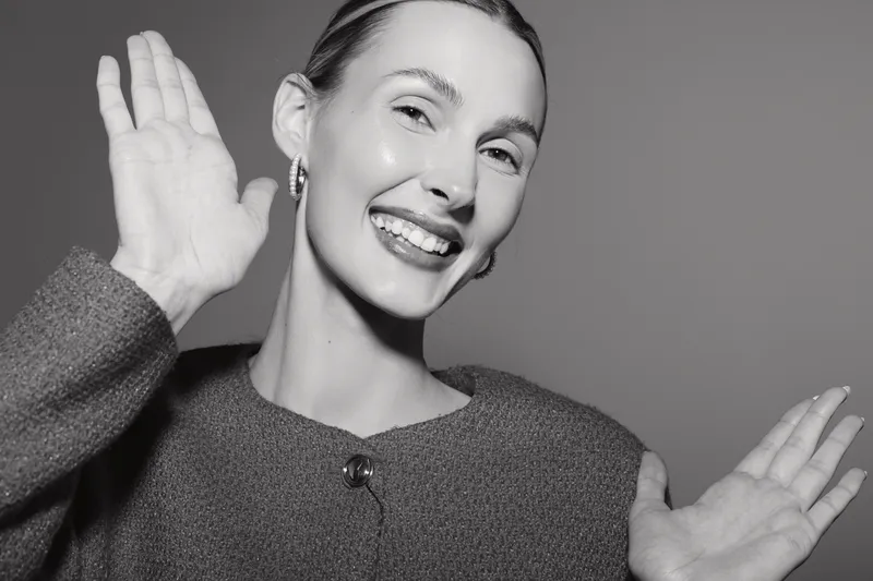 Young Caucasian woman with fair skin smiling and waving hands in a studio with plain background.
