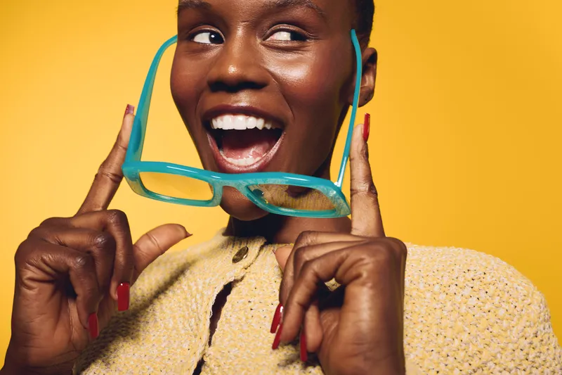 Happy black woman with dark skin holds turquoise glasses near face, smiling widely against yellow background. Concept of joy, fashion, beauty, and positive emotions in studio portrait.