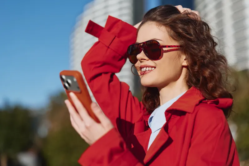 Young woman in a bright red coat smiles while scrolling on her smartphone outdoors, wearing sunglasses and enjoying a sunny day with a clear blue sky behind.