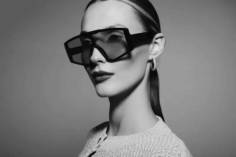 Stylish Caucasian woman with pale skin wearing oversized glasses and earrings looks confident in a black and white studio portrait.
