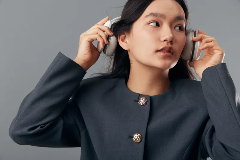 Woman, headphones, listening and fashion with gray jacket against background. Young adult female, audio device and lifestyle with casual style for music and sound entertainment.
