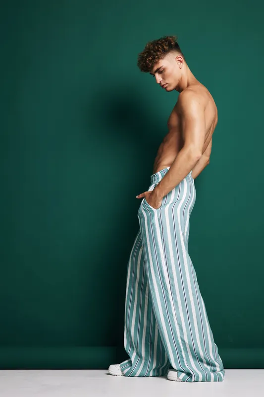 Young man with curly hair wearing striped wide leg pants and white sneakers standing sideways against green background with hands in pockets and serious expression.