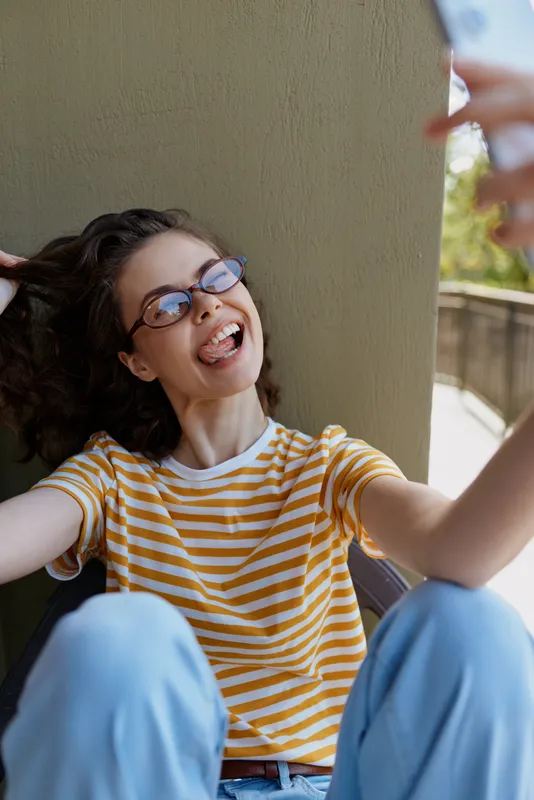 A woman taking a selfie outdoors, wearing striped yellow shirt and jeans, smiling through glasses. A vivid lifestyle moment capturing casual confidence and self expression.