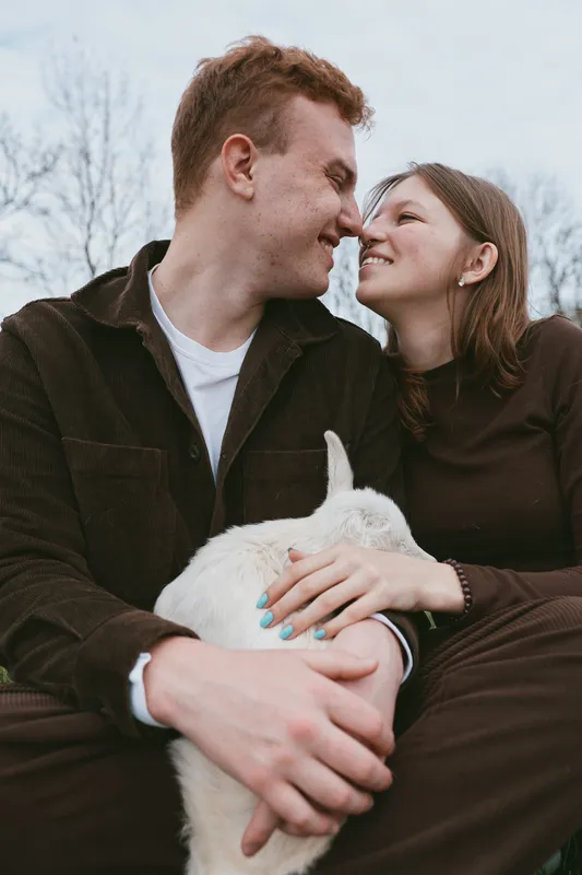Warm lifestyle scene shows a couple outdoors with a goat kid, sharing a loving moment, smiling at each other as the animal rests in their arms, creating a cozy, intimate family vibe.