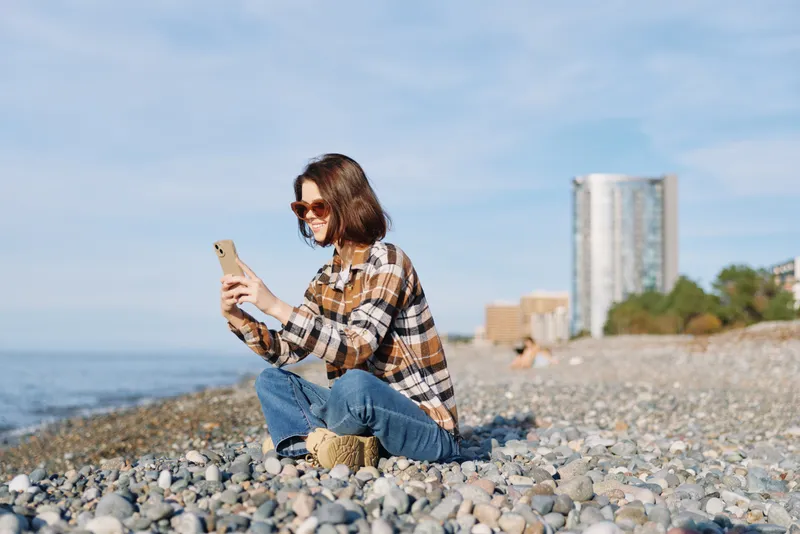 Casual woman on a pebble beach using a smartphone by the sea with distant buildings, enjoying the outdoors and coastal lifestyle, connectivity and relaxed moment
