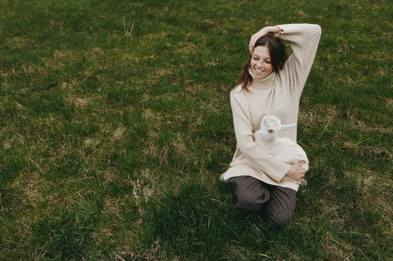Outdoor lifestyle moment with a smiling woman kneeling in grass, holding a goat kid, cozy knit sweater, warm sunlight, natural colors, peaceful countryside vibe, candid connection